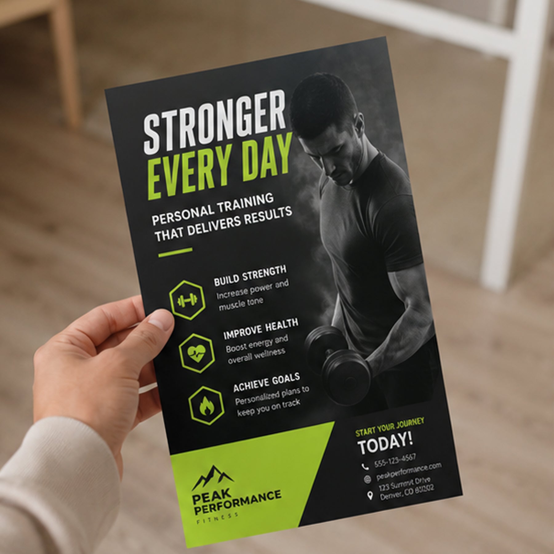 A hand holds a printed fitness flyer or poster in a real indoor setting. The flyer has a black background with a grayscale photo of a man lifting a dumbbell, large white and lime-green text reading 'STRONGER EVERY DAY,' and a bright lime-green section at the bottom with the Peak Performance Fitness logo and contact details.